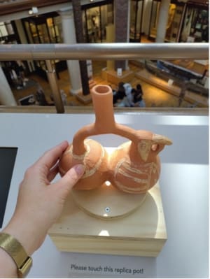 A hand touching the replica moche pot. The pot is made of two connected circles and in one of the ends it has the face of a parrot. Details are painted with white paint.