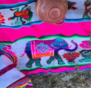 Bright pink synthetic textiles laid on the ground in Huilloc Alto, bordered in green and blue. The woven designs include flowers and non-native animals, including an elephant and a lion. A copper vessel rests on the textile, and part of an artisan’s multicolored clothing appears at the lower left.