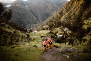 Two artisans wearing traditional red, orange, and black clothing walk along a mountain path in Huilloc Alto. Green fields, scattered trees, steep slopes, and the valley extend behind them.
