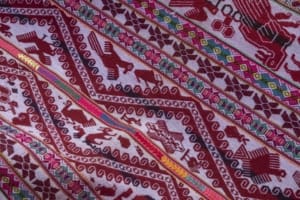 Close-up of a red and white woven lliqlla arranged in diagonal bands. The textile includes motifs of birds, horses, flowers, a heart, and other animals, separated by geometric borders and narrow multicolored stripes.