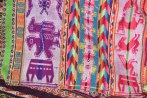 Close-up of a brightly colored woven textile with vertical bands in pink, green, orange, purple, and white. In the upper pink and purple sections, boat motifs appear as basket-like geometric forms. Other bands include animals and intricate multicolored patterns.