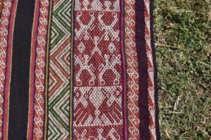Close-up of a woven textile laid on grass. In the central red-and-white panel, a human figure appears at the center of a symmetrical composition, with four horse-like figures extending outward. The panel is bordered by black, green, red, white, and multicolored geometric bands.