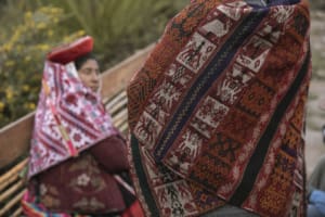 Rear view of an artisan wearing a richly patterned lliqlla in red, white, orange, black, and purple. Several woven panels show horses with human riders on their backs. Another artisan wearing a red-and-white textile appears blurred in the background.