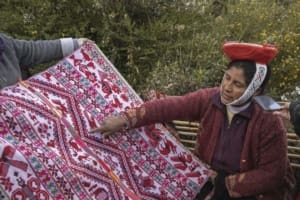 Two artisans wearing traditional red, orange, and black clothing walk along a mountain path in Huilloc Alto. Green fields, scattered trees, steep slopes, and the valley extend behind them.