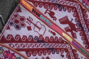 Close-up of a red and white woven lliqlla with geometric borders and figurative motifs, including birds, flowers, animals, and a guitar, crossed by narrow bands of bright colors.