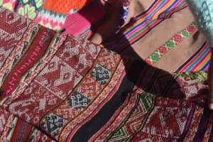Close-up of several layered woven textiles in red, white, black, orange, green, and pink. A central red-and-black motif is divided into upper and lower sections representing a flower and its root. To the left, bird motifs appear arranged in pairs within a red-and-white panel.